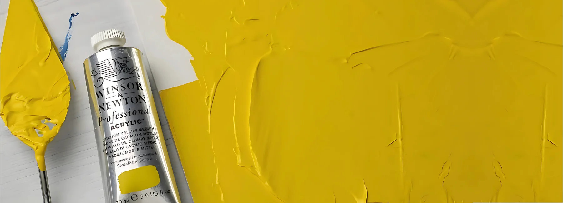 winsor-and-newton-professional-acrylic-paint-unleashing-creativity. on the left hand side of the image is a palette knife covered in yellow acrylic paint. to the right of it is a tube of proffessional acrylic paint it is silver with a white cap in a tube. the rest of the image on the right of them is a canvas panel that has been thickly covered in the yellow paint.