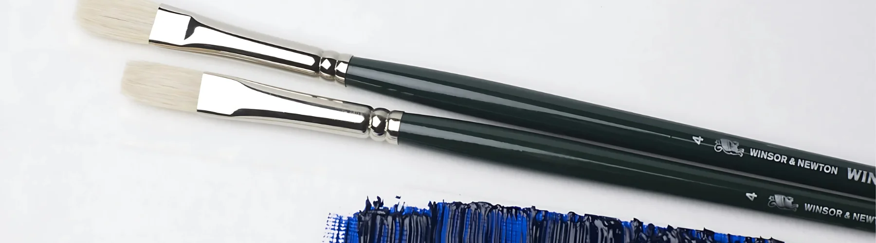 winsor-and-newton-winton-brushes-making-masterpieces. in the center of the image are two winton brushes. they are hair facing the left. they have long green handles with silver ferrules and white hairs. infront of them is oil paint in a deep blue colour that has been painted roughly in a small patch on the bottom right hand side of the image. on a white background