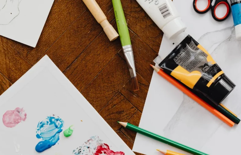 art-paper-supplies-a-discovery. the image is a birds eye view of a desk with an assortment of art supplies on it. paints, brushes, pens, scissors and paper that has paint on it. on a wooden table