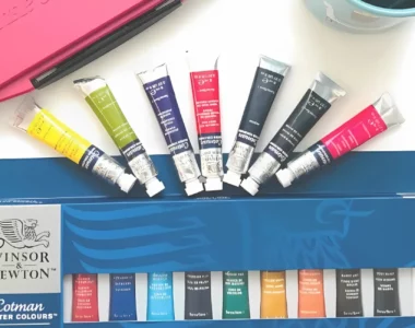 cotman-watercolour-paints-the-winsor-newton-promise. the image is a birds eye view of a set of cotman tubes still in there box with the individual tubes in a half circle shape above it and sitting on watercolour paper that has paint on it. with a pink storage case behind it and a glass with water in at the top