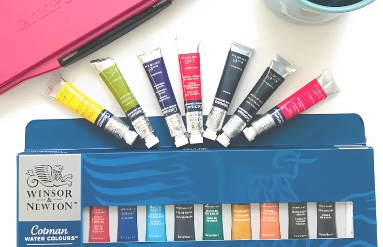 cotman-watercolour-paints-the-winsor-newton-promise. the image is a birds eye view of a set of cotman tubes still in there box with the individual tubes in a half circle shape above it and sitting on watercolour paper that has paint on it. with a pink storage case behind it and a glass with water in at the top