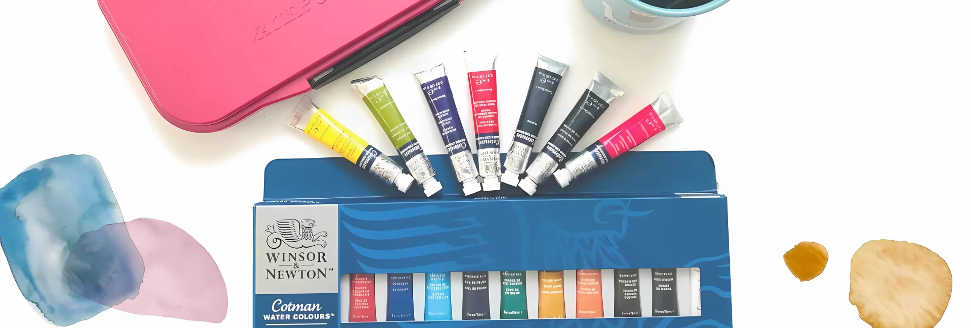 cotman-watercolour-paints-the-winsor-newton-promise. the image is a birds eye view of a set of cotman tubes still in there box with the individual tubes in a half circle shape above it and sitting on watercolour paper that has paint on it. with a pink storage case behind it and a glass with water in at the top