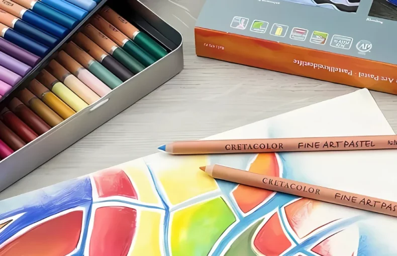 cretacolor-pastel-pencils-artists-essentials. the image is a close up of a set of cretacolour pastel pencils that have been opened and are still in the tin. the tin is fanned out to see both the layeers of pencils inside it. infront is a paper that has an image drawn on it using the pastel pencils in bright colours. there are two pencils sitting on the paper. in a orange and blue colours. on a tabletop that is light in colour