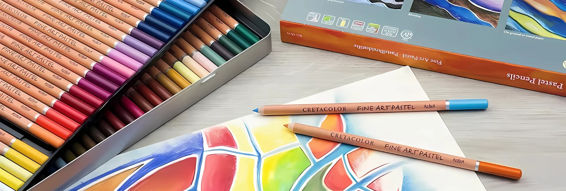 cretacolor-pastel-pencils-artists-essentials. the image is a close up of a set of cretacolour pastel pencils that have been opened and are still in the tin. the tin is fanned out to see both the layeers of pencils inside it. infront is a paper that has an image drawn on it using the pastel pencils in bright colours. there are two pencils sitting on the paper. in a orange and blue colours. on a tabletop that is light in colour