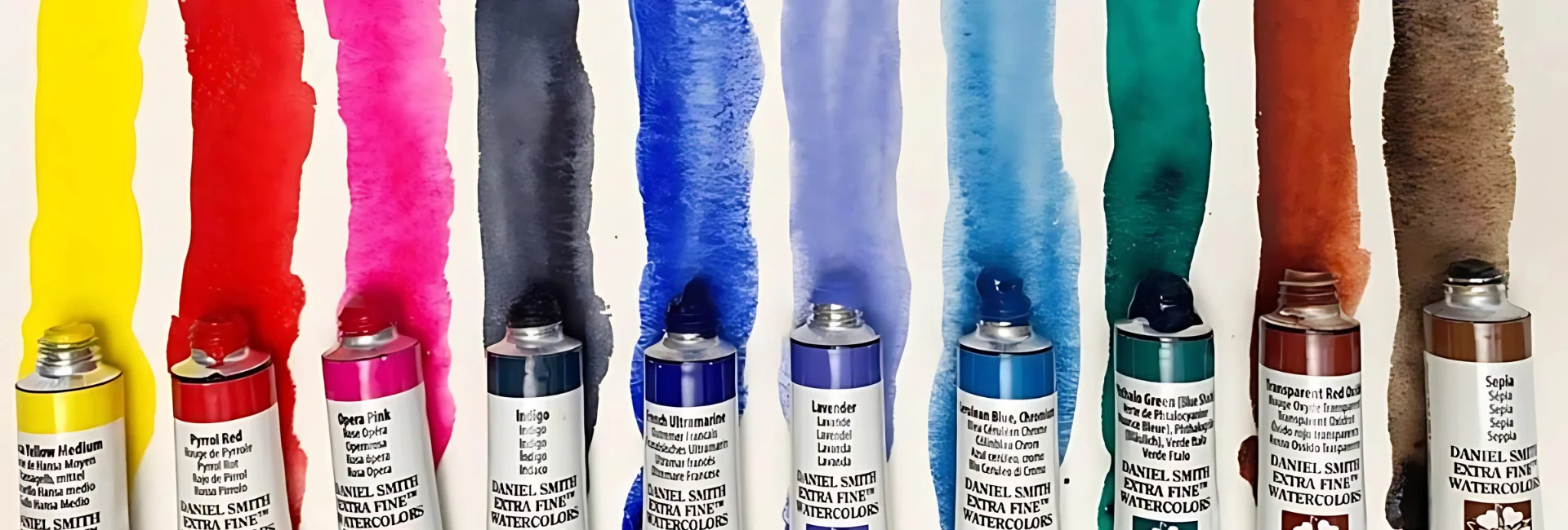 daniel-smith-watercolour-brushes-at-the-ready. the image is a close up of a rainbow selection of daniel smith watercolour tubes that are open and a streak of colour has been painted underneath and in front of them in the same colour that is in the tube. on watercolour paper. the colours are rich and vibrant looking