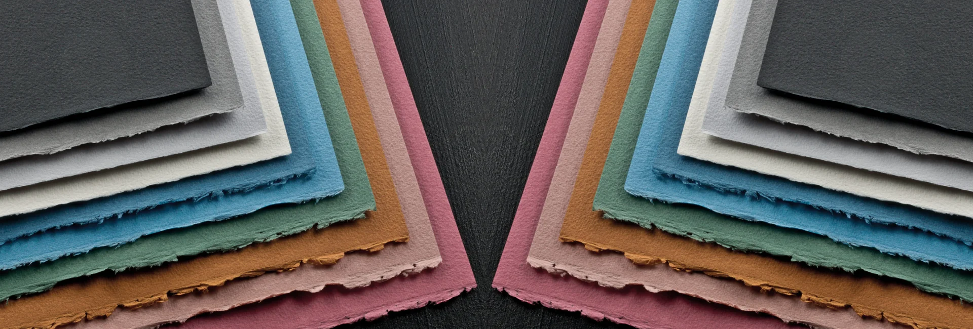 fabriano-cromia-paper-delve-into-artistry. the image is of a mirror image of the same picture of various colours of the cromia paper fanned out to show the different colours and coming from the outer edge going in. on a dark wood table top