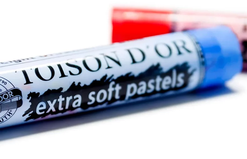 koh-i-noor-toison-dor-soft-pastels, the image is a close up of two of the extra soft pastels. the left hand one is blue and the right hand one is red. they are taking over most of the image. the name is cleasrly visable on the blue and the red pastel is out of focus. they are on a white background