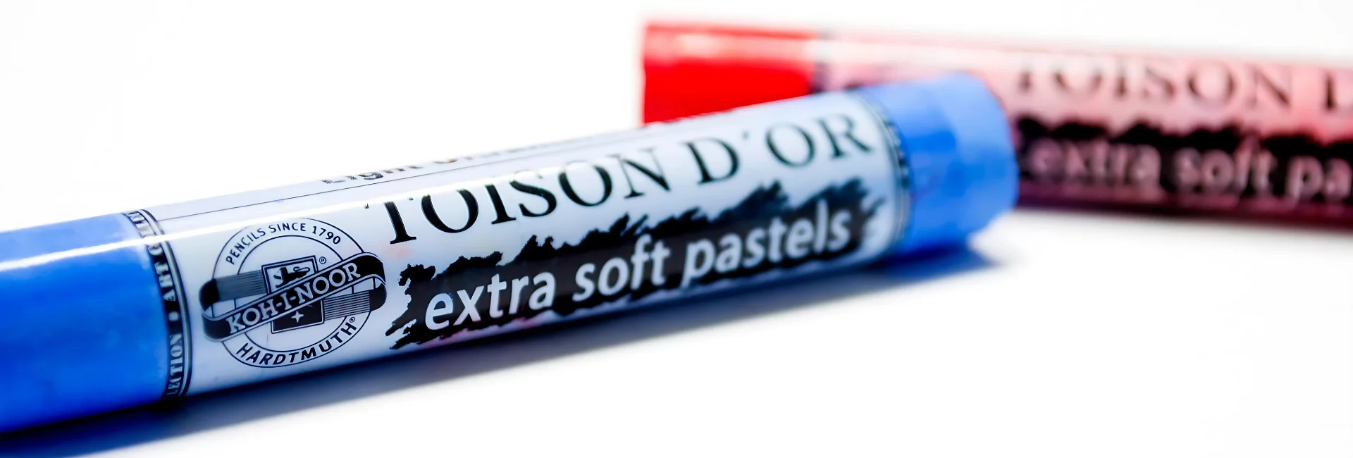 koh-i-noor-toison-dor-soft-pastels, the image is a close up of two of the extra soft pastels. the left hand one is blue and the right hand one is red. they are taking over most of the image. the name is cleasrly visable on the blue and the red pastel is out of focus. they are on a white background