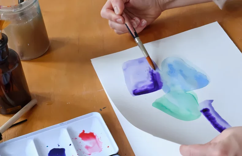 lukas-aqua-studio-artists-watercolours. the image is a close up of a woman painting a watercolour painting. she is sitting at a desk and has a paintbrush in her hand and is applying blue watercolour paint to paper. around her on the desk are various painting suppllies. a palette with paint on it and a jar of brushes and water to dip her brush in. the background is a blurred visual of the room she is in.