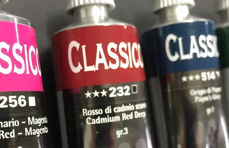 maimeri-classico-oil-paint-a-pigment-pleasure is shown in a line across the center of the image. there is a pink, red, blue, green and yellow tube. they are lying off center and the name is visable. on a grey background