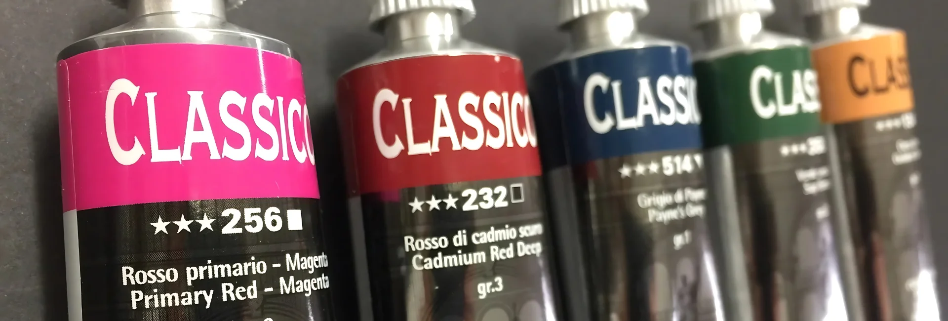 maimeri-classico-oil-paint-a-pigment-pleasure is shown in a line across the center of the image. there is a pink, red, blue, green and yellow tube. they are lying off center and the name is visable. on a grey background
