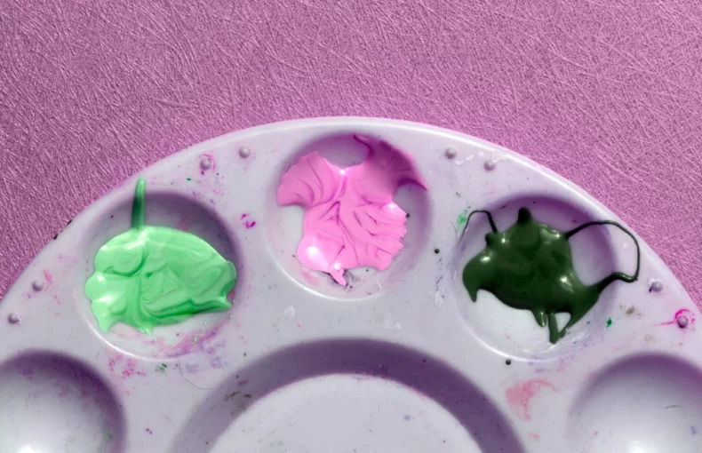 paint-palettes-an-artists-staple. the image is a close up of a single plastic palette. it is round and you can only see the top of it. the top three wells have different coloured paint in them. on a pink background