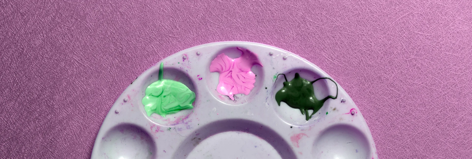 paint-palettes-an-artists-staple. the image is a close up of a single plastic palette. it is round and you can only see the top of it. the top three wells have different coloured paint in them. on a pink background