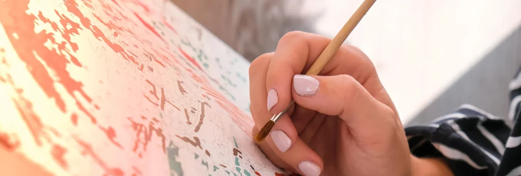 the image is a close up of a paint by numbers canvas and a womans hand with painted pink nails holding a painbrush and acting as though she is painting on it. the background is a blur of the room behind her.