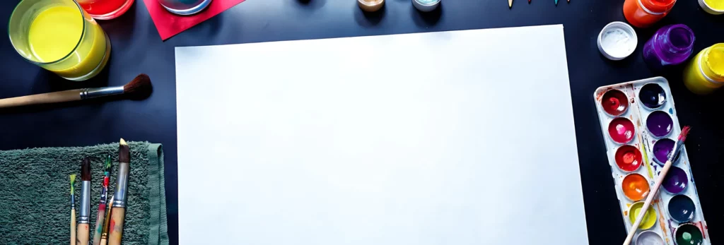 the image is a birds eye view of a table with painting supplies on it. in the center is a sheet of watercolour paper that is blank and around it are paint sets, brushes, sponges, paints, rollers and dipping water for the brushes. on a dark blue tabletop. the light is coming from the top of the image.