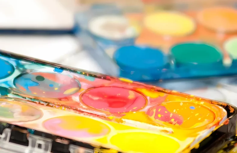 paint-sets-brushing-up-on-inspiration. in the center of the image is a close up of two watercolour paint pan sets. the one in the front has been used and there is paint all over it. the background one is still new and sealed with a clear plastic container. they are sitting on top of watercolour paper that has paint splotches on it.
