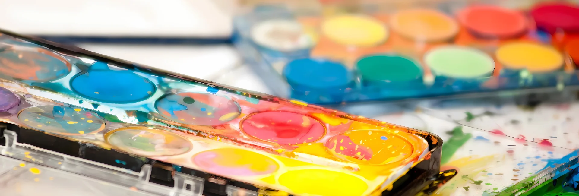 paint-sets-brushing-up-on-inspiration. in the center of the image is a close up of two watercolour paint pan sets. the one in the front has been used and there is paint all over it. the background one is still new and sealed with a clear plastic container. they are sitting on top of watercolour paper that has paint splotches on it.
