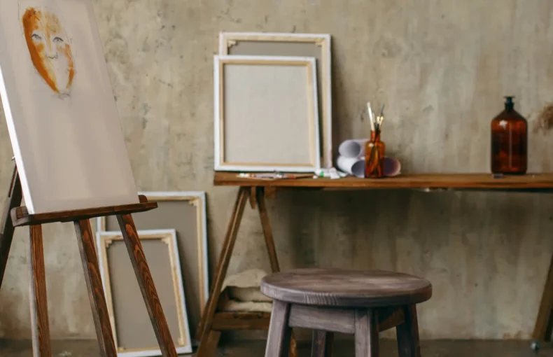 painting-tools-for-aspiring-artists. the image shows an art room with easels in it on the left and canvas on the easel and leaning against walls. there is a table with various art supplies on it and chairs scattered around the room. the wall behind is a dirty grey colour
