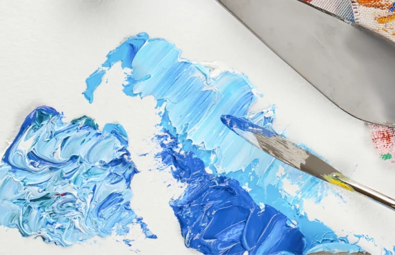 palette-knives-pizzazz-elevate-your-art. the image shows two palette knives coming in from the right hand side of the image. they have blue and white paint on the and they are mixing the paint on a surface. behind them are cloths with paint on them