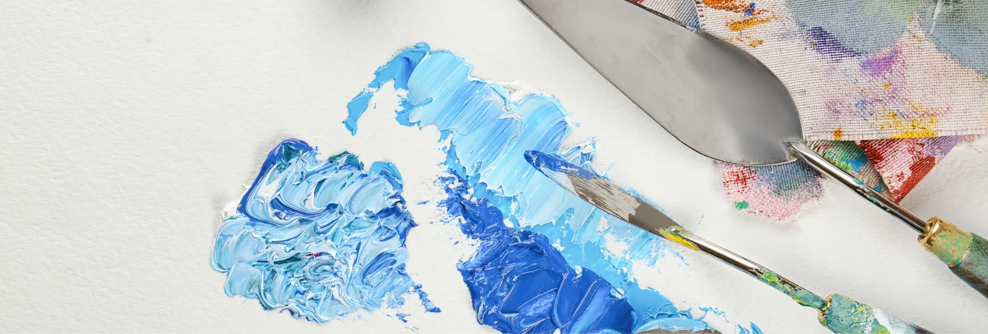 palette-knives-pizzazz-elevate-your-art. the image shows two palette knives coming in from the right hand side of the image. they have blue and white paint on the and they are mixing the paint on a surface. behind them are cloths with paint on them