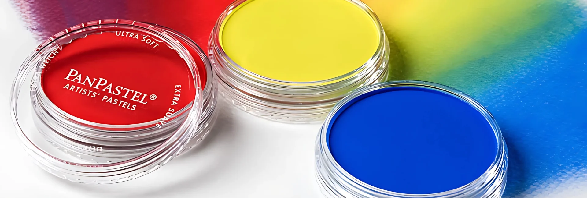 panpastel-sets-unleash-your-creativity. the image is a close up of three pots of pan pastels. in red,yellow and blue. the red still has the lid half on and the yellow and blue have no lid. behind them are the blended colours of the three. on a white surface.