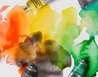 professional-watercolour-paint-by-winsor-newton. the image is of various proffessional watercolour tubes scattered across a watercolour paper in different colours with their lids off. the paper is a rainbow blot wash of the watercolour paints. its very vibrant and bright