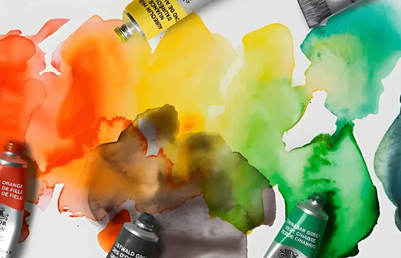 professional-watercolour-paint-by-winsor-newton. the image is of various proffessional watercolour tubes scattered across a watercolour paper in different colours with their lids off. the paper is a rainbow blot wash of the watercolour paints. its very vibrant and bright