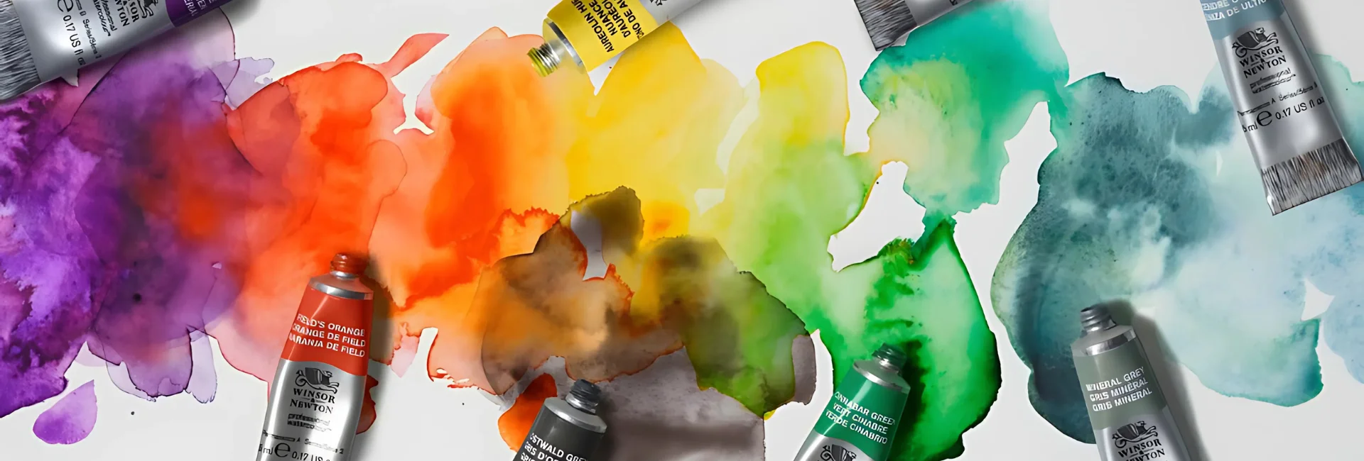 professional-watercolour-paint-by-winsor-newton. the image is of various proffessional watercolour tubes scattered across a watercolour paper in different colours with their lids off. the paper is a rainbow blot wash of the watercolour paints. its very vibrant and bright
