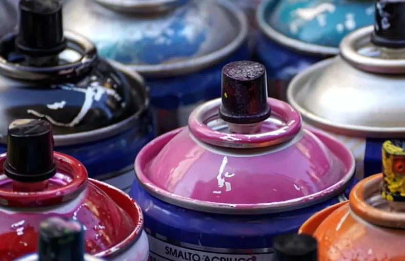 spray-paints-discover-the-power. in the center of the image is a close up of the spray nozzles of the spray paint cans. they are a variety of different colours. reds, yellows, blues, greens, orange and pink. there is reidue of the spray paint on the black nozzles and the tops of the cans have paint on them.they are packed tightly in the image.