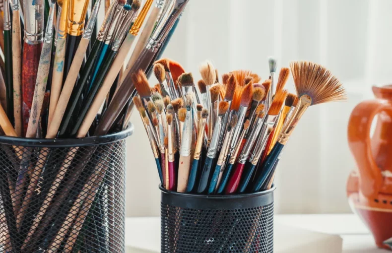 storage-solutions-a-painting-supplies-best-friend. the image shows various holders on the table storing paint brushes. there are easela in the background holding up the art works.on a table and the background is a blurred visual of the room behind