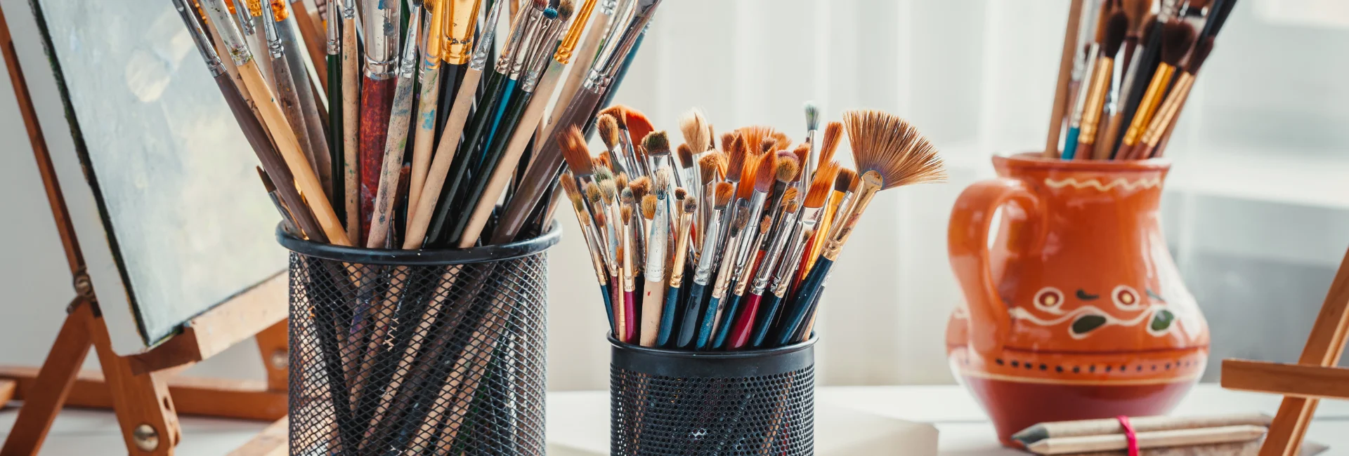 storage-solutions-a-painting-supplies-best-friend. the image shows various holders on the table storing paint brushes. there are easela in the background holding up the art works.on a table and the background is a blurred visual of the room behind