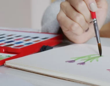 watercolour-paper-unleash-your-creativity. the image shows a woman sitting at a table and holding a red paintbrush and painting flowers on a watercolour paper. next to her is a red palette with watercolour paint in it. the background is a blurred visual of the room behind her