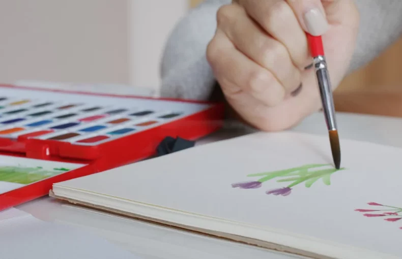 watercolour-paper-unleash-your-creativity. the image shows a woman sitting at a table and holding a red paintbrush and painting flowers on a watercolour paper. next to her is a red palette with watercolour paint in it. the background is a blurred visual of the room behind her