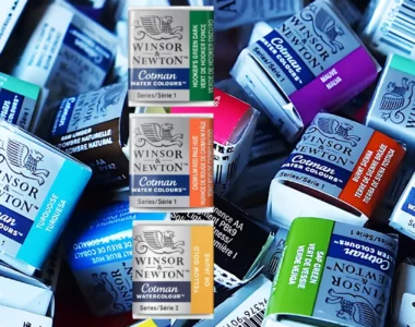 winsor-and-newton-cotman-watercolour-half-pans-colour-your-world. the image shows two duplicated images of a pile of cotman pans and in a big pile and in a variety of colours. in the center are three induvidual pans in a row. a green, orange and yellow.