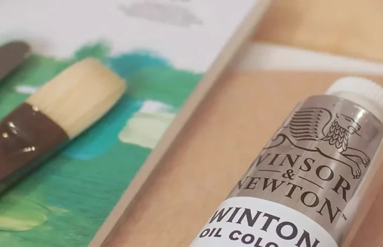 winsor-and-newton-oil-colours-the-artisans-choice. in the center of the image is a clise up of two white winton oil paints. you can just see the name and top of the tube. to the left of it is a variety of palette knives and a paintbrush sitting on a canvas that has green painted on it.