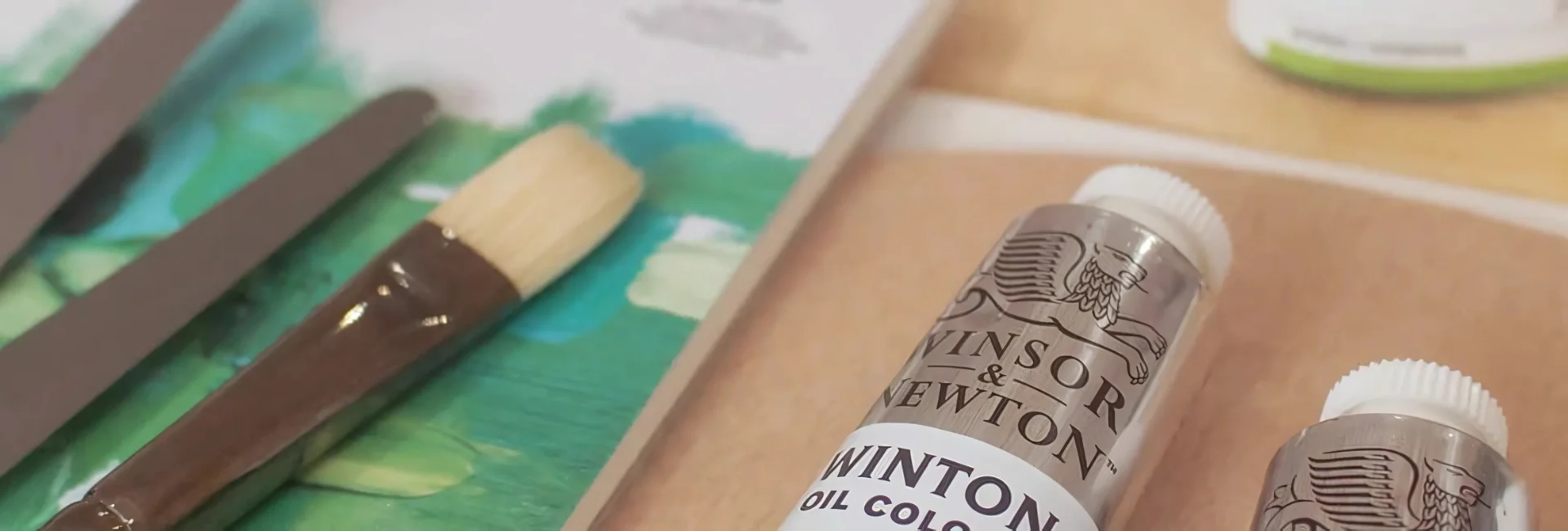 winsor-and-newton-oil-colours-the-artisans-choice. in the center of the image is a clise up of two white winton oil paints. you can just see the name and top of the tube. to the left of it is a variety of palette knives and a paintbrush sitting on a canvas that has green painted on it.