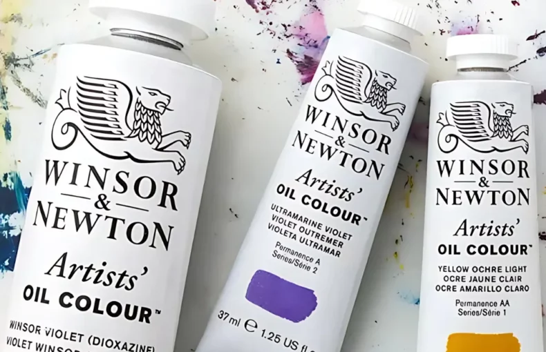 winsor-and-newton-oil-paint-discovering-the-artistic-journey. in the center of the image are three tubes of the winsaor and newton artists oil paint. they are lying next to eachother on a canvas sheet that has various of colours of paint on it. the tubes are white and have white caps and black writing on them. they are in yellow, purple and blue colours. on a table top that has paint in different colours smeared on it.