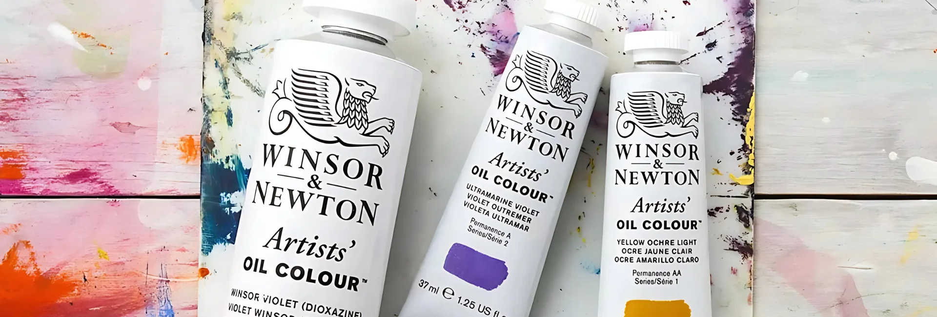 winsor-and-newton-oil-paint-discovering-the-artistic-journey. in the center of the image are three tubes of the winsaor and newton artists oil paint. they are lying next to eachother on a canvas sheet that has various of colours of paint on it. the tubes are white and have white caps and black writing on them. they are in yellow, purple and blue colours. on a table top that has paint in different colours smeared on it.