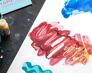 yupo-paper-where-art-magic-collide-1. the image is of a sheet of yupo on a table with various art mediums around it. water in a glass, paintbrushes, watercolour paint, acrylic paint, alcohol inks all being used on the yupo to show what mediums work on it and how they work on it