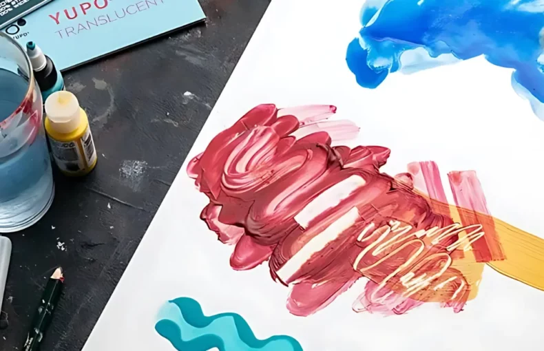 yupo-paper-where-art-magic-collide-1. the image is of a sheet of yupo on a table with various art mediums around it. water in a glass, paintbrushes, watercolour paint, acrylic paint, alcohol inks all being used on the yupo to show what mediums work on it and how they work on it